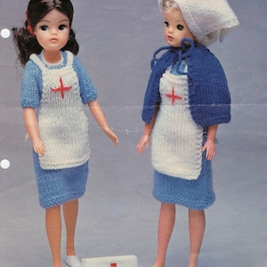 May include: Two dolls dressed as nurses, wearing white and blue knitted dresses with red crosses. One doll is wearing a white knitted hat with a red cross. The dolls are standing next to a red cross first aid kit.