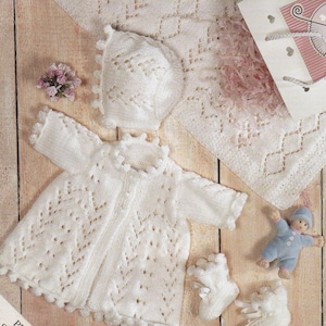 Puede incluir: Un conjunto de bebé tejido en blanco, que incluye una chaqueta, gorro y patucos, con una manta a juego. El conjunto se exhibe sobre una superficie de madera con una pequeña muñeca y una bolsa de regalo. La chaqueta y los patucos tienen un ribete de pompón.