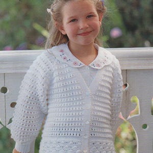 May include: A young girl in a white, crocheted cardigan with a V-neck and button closure. The cardigan features a textured pattern and long sleeves. The collar of the shirt underneath is white with pink floral embroidery. The girl is smiling.