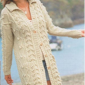 Op de afbeelding: Crèmekleurig, lange cardigan met een textuur gebreid ontwerp. Voorzien van een kraag, knoopsluiting aan de voorkant en twee voorzakken. De trui wordt gedragen met een witte top en een spijkerbroek. Er wordt ook een bruine hoed gedragen.