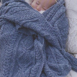 Puede incluir: Un niño pequeño durmiendo profundamente envuelto en una manta de punto azul con flecos. La manta es texturizada y tiene un patrón de punto de cable. El niño está acostado en una cama blanca con una sábana con estampado floral.