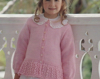 May include: A light pink knitted cardigan with a white lace collar and button closure. The cardigan features a decorative knitted pattern at the hem. The girl is wearing a white dress. The cardigan is a children's clothing item.