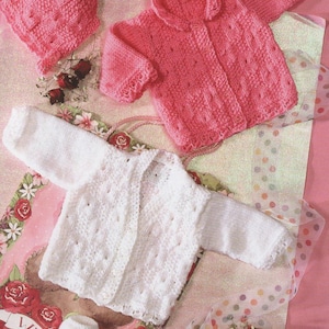 Könnte beinhalten: Eine Sammlung handgestrickter Babykleidung in zartem Rosa und Weiß. Das Set beinhaltet eine Mütze, eine Jacke mit Kragen, eine weiße Strickjacke und ein Paar Babyschuhe. Die Kleidungsstücke sind auf einem rosa Blumengrund drapiert.
