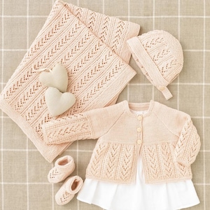 Puede incluir: Un conjunto de ropa de bebé de punto en color rosa pálido, que incluye un cárdigan, un gorro, botines y una manta. Todos los artículos están hechos con un delicado patrón de encaje.