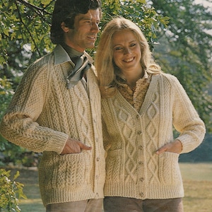 May include: A pair of cream-colored cable knit cardigans with pockets and button closures. The cardigans are worn by two people standing in a grassy field.