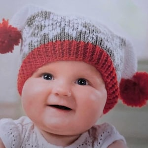May include: A baby wearing a white and gray knitted hat with red pom-poms and a red and white striped band.