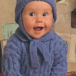 Könnte beinhalten: Ein Baby trägt einen blauen Strickpullover mit Zopfmuster und eine passende blaue Strickmütze mit Bommel.