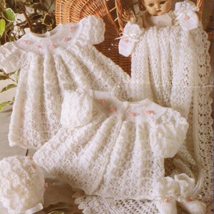 May include: A collection of white, crocheted baby garments, featuring a dress, bonnet, booties, and a blanket. The dress and bonnet are decorated with tiny pink flowers. The set is presented with a doll in a wicker basket, giving a vintage feel.