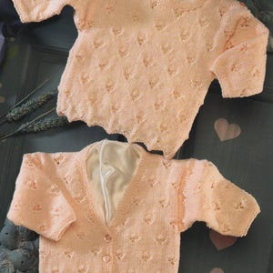May include: Two light peach-colored baby sweaters. One is a crew neck with three buttons on the shoulder, and the other is a cardigan with a V-neck and button closure. Both sweaters have a textured pattern.