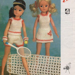 May include: Two dolls in knitted tennis outfits, one with shorts and the other with a dress. Both outfits are white with red trim. One doll holds a tennis racket and a yellow ball. The background is a light blue.