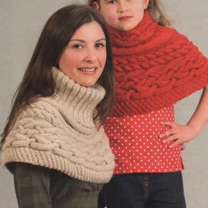 May include: Two people wearing knitted cowl neck sweaters. One is a light beige color, and the other is a bright red. The red sweater is worn over a red polka dot shirt. The sweaters have a cable knit design.