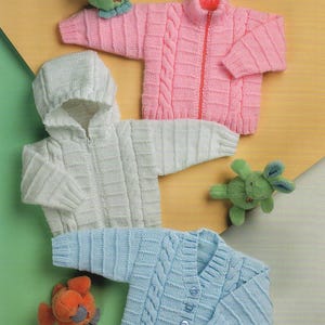 May include: Three knitted baby cardigans in pink, white and blue. The cardigans feature a cable knit pattern and are zipped or buttoned. The cardigans are displayed on a light brown surface with soft toys.