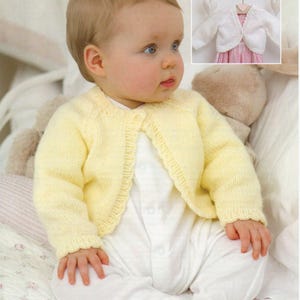 May include: A baby wearing a pale yellow knitted cardigan with a scalloped edge, over a white onesie. The cardigan has long sleeves and a button closure. A smaller image shows a white bolero jacket on a hanger.