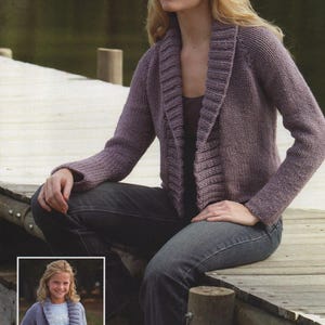 May include: A woman wearing a purple knitted cardigan with a wide collar, sitting on a wooden jetty. She is wearing a hat and jeans.