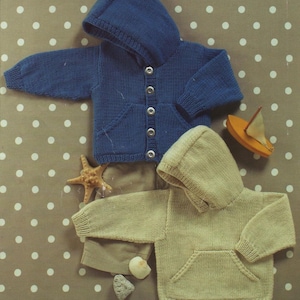 May include: Two knitted baby hoodies, one in blue and one in beige, with hoods and front pockets. The blue hoodie has silver buttons. A starfish, toy sailboat, and seashells are arranged nearby.