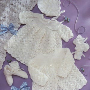 Puede incluir: Un conjunto de ropa de bebé de punto blanco, que incluye un vestido, una rebeca, un gorro, patucos y una manta. Los artículos están dispuestos sobre una superficie morada y acentuados con cintas azules y blancas.