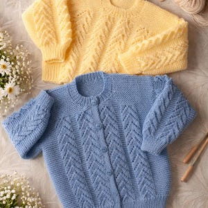 Könnte beinhalten: Zwei Strickpullover, einer gelb und einer hellblau, werden gezeigt. Der gelbe Pullover ist ein Pullover, während der blaue eine Strickjacke mit Knöpfen ist. Beide haben ein strukturiertes Zickzackmuster. Garn, Holzstricknadeln und Knöpfe sind ebenfalls sichtbar.