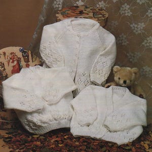 May include: Three white knitted baby sweaters with button closures and decorative openwork patterns are displayed. A teddy bear and two decorative boxes are in the background. The sweaters are arranged on a bed of dried leaves.
