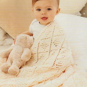 Könnte beinhalten: Ein Baby ist in eine weiße Häkeldecke mit einem Teddybären in den Armen gewickelt. Die Decke hat ein Spitzenmuster und ist aus weichem Garn gefertigt.