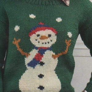 Peut inclure: Un pull en tricot vert avec un motif de bonhomme de neige blanc. Le bonhomme de neige porte une écharpe rouge et bleue et des boutons noirs. Le pull a un col à carreaux blancs et noirs.