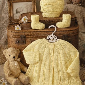 May include: A collection of handmade baby items in soft yellow, including a knitted cardigan, hat, and booties. A teddy bear and a vintage doll are also present, creating a nostalgic, cozy scene. The cardigan is on a white hanger.
