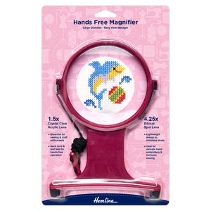 May include: A burgundy hands-free magnifier for crafts and sewing, with a 1.5x acrylic lens and a 4.25x bifocal spot lens. It includes a neck cord and rest feet for hands-free use. The image shows a cross-stitch design. The text reads 'Hands Free Magnifier'.