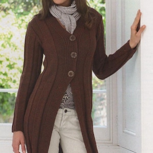 May include: A long, brown knit cardigan with three large buttons, worn over a patterned top and light-colored pants. The person is also wearing a gray hat and scarf. The cardigan has a ribbed texture.