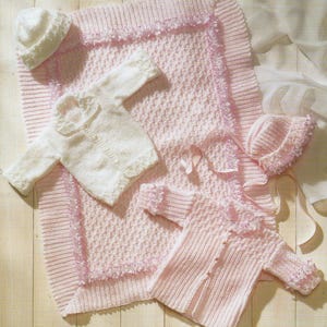 May include: A collection of baby knitwear in soft pink and white. The set includes a blanket, two sweaters, and two hats. The blanket has a textured pattern and a ruffled border. The sweaters have button closures and ruffled collars. The hats are also knitted.