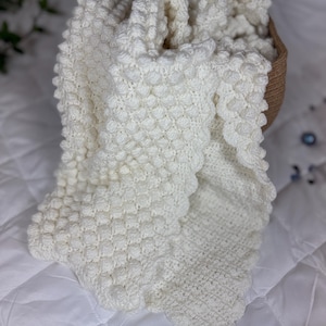 Op de afbeelding: Een wit gebreid babydekentje met een textuur, een bolletjespatroon. Het dekentje is gevouwen en ligt in een geweven mand.