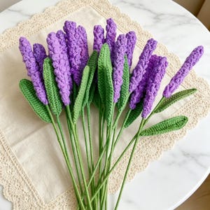 Könnte beinhalten: Ein Strauß aus handgefertigten gehäkelten Lavendelblüten mit leuchtend violetten, strukturierten Blütenständen und langen grünen Blättern an dünnen Stielen. Jede Blume ist sorgfältig mit Garn gehäkelt und zeigt eine detaillierte botanische Struktur mit mehreren Stielen, die zu einem Bündel zusammengefasst sind.