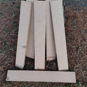 May include: A collection of unfinished, light-colored wooden planks, arranged against a tree trunk and on a bed of pine needles. The planks vary in length and width, showcasing the natural wood grain. Ideal for woodworking projects.
