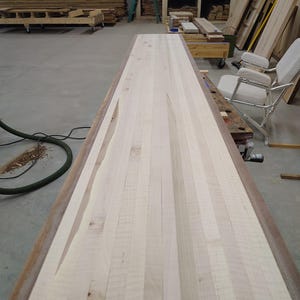 Puede incluir: Una encimera de madera larga y de color claro con un borde marrón oscuro. La encimera está hecha de tiras verticales de madera, mostrando la veta natural. La madera tiene algunas imperfecciones y nudos, lo que le da un aspecto rústico. La encimera está en un taller.