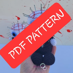 Puede incluir: Un letrero rojo y blanco con el texto "PDF PATTERN" que cubre una muñeca de crochet con una cabeza blanca cubierta de clavos y un cuerpo negro. La muñeca está siendo sostenida por una mano.