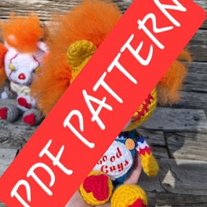 May include: A crochet doll with bright orange hair, a yellow head, and blue overalls featuring a white patch with the red text Good Guys. The doll has red accents on its feet and a red stitched mouth. A second smaller crochet doll is visible behind.