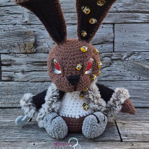 May include: A brown crochet rabbit plush toy with large red eyes, wearing a black and faux fur cape. The rabbit features bee-patterned fabric on the inner ears and several small bee embellishments on its head and chest. The text Mamas Creations is visible.