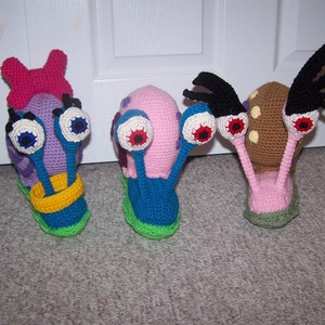 Op de afbeelding: Drie gehaakte slakken knuffels met grote ogen. De slakken zijn roze, paars en bruin met groene en blauwe accenten.