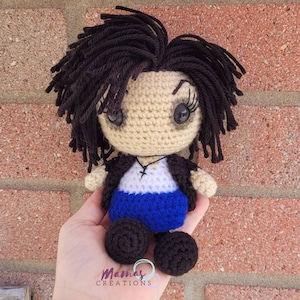 May include: Crocheted doll with black hair, a white shirt, a blue skirt, and black shoes. The doll is sitting on a hand.