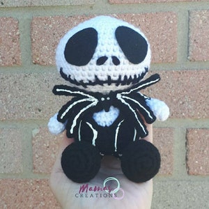 May include: A crocheted plush toy of Jack Skellington from the Nightmare Before Christmas. The toy is white with black details and has a black bow tie. The toy is sitting on a hand.