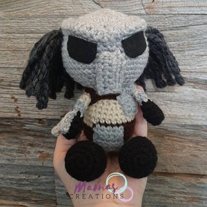 May include: Crocheted plush toy of a Predator character. The toy is grey with black eyes and dreadlocks. It is wearing a brown and beige vest and has black feet.