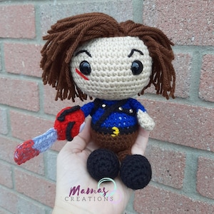 May include: Crocheted plush toy of a character with brown hair, a blue shirt, brown pants, and a red and blue chainsaw. The toy has black eyes and a red scar on its face.