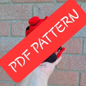 Op de afbeelding: Een handgemaakte gehaakte lieveheersbeestje, voornamelijk rood met zwarte vlekken en details, vastgehouden in een hand. Het lieveheersbeestje heeft een afgerond lichaam en kleine antennes. Een rode banner met de tekst "PDF PATTERN" is over de afbeelding geplaatst.