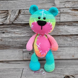 Può includere: Un orsacchiotto di peluche all'uncinetto arcobaleno con un cuore rosa sul petto. L'orso ha un naso rosa cucito e occhi neri.