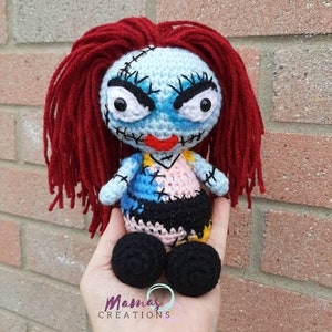 May include: A crocheted doll with red hair, blue and pink patchwork clothing, and black eyes. The doll has a stitched-on smile and is holding a red heart. The doll is sitting on a hand.