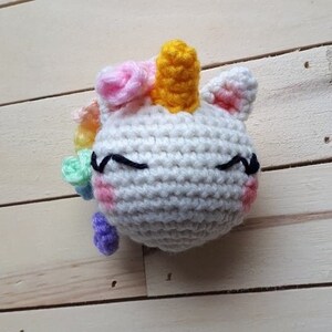May include: A white crocheted unicorn with a yellow horn, pink and green flowers, and closed eyes. The unicorn is on a wooden background.