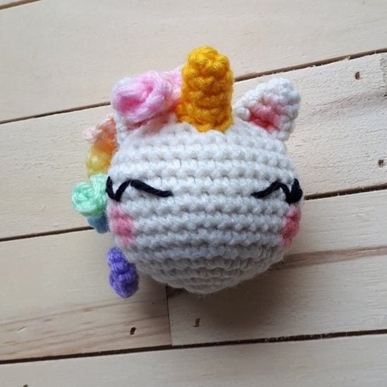 May include: A white crocheted unicorn with a yellow horn, pink and green flowers, and closed eyes. The unicorn is on a wooden background.