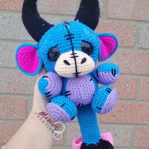May include: A blue crocheted plush toy with black horns, pink ears, and a long tail. The toy has a stitched-up appearance with purple accents and large black eyes.
