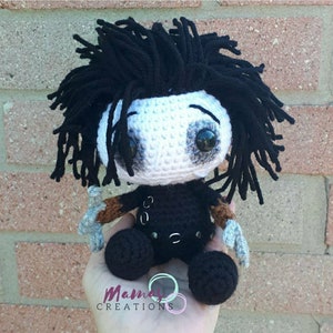 May include: Crocheted plush toy of a character with black hair, white face, and black clothing. The toy is holding two silver knives in its hands. The toy is sitting on a hand.