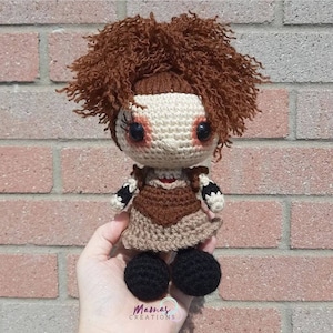 May include: Crocheted doll with brown hair, a brown dress, and black shoes. The doll has a white face with black eyes and red blush.