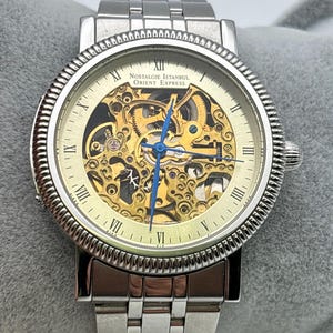 May include: A silver-toned wristwatch with a skeleton dial, showcasing the inner workings of the mechanism. The watch features Roman numeral hour markers, blue hands, and the text "NOSTALGIE ISTANBUL ORIENT EXPRESS" on the face. The watchband is also silver-toned.