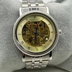 May include: Silver-toned wristwatch with a skeleton dial, showcasing the intricate inner workings. The watch features Roman numeral hour markers and the text "Nostalgie Istanbul Orient Express" on the face. The watch band is made of linked metal.